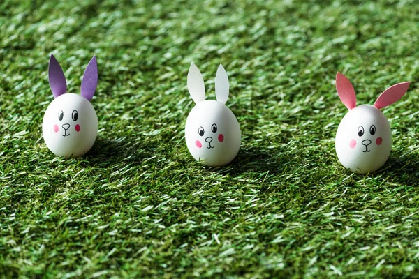 Foyer sélectif des œufs de poulet avec des visages de lapin dessinés et des oreilles en papier sur la surface de l'herbe verte — Photo de stock