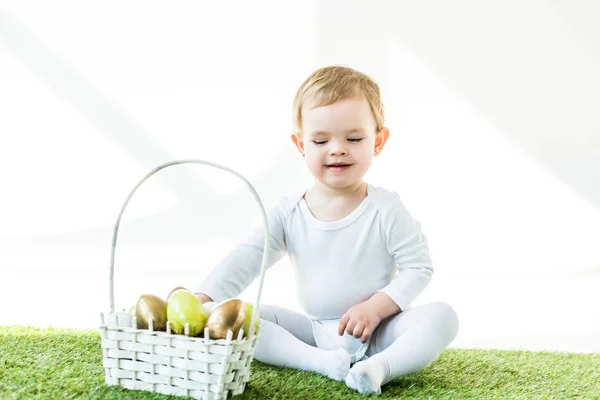 Cute smiling baby sitting near straw basket with Easter eggs isolated on white — Stock Photo