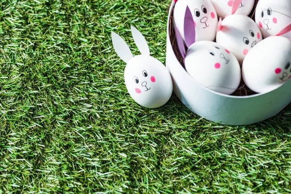 Ovos de galinha com caras de coelho engraçado e orelhas de papel na superfície de grama verde — Fotografia de Stock