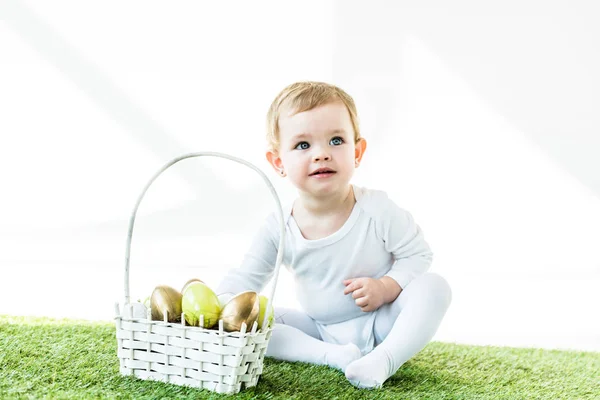 Adorável bebê sorridente sentado perto da cesta de palha com ovos de Páscoa isolados em branco — Fotografia de Stock