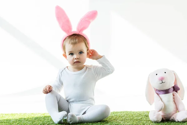 Bonito criança no coelho orelhas headband sentado perto de brinquedo coelho isolado no branco — Fotografia de Stock