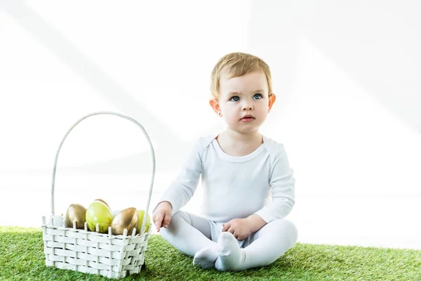 Lindo bebé sentado cerca de la cesta de paja con huevos de Pascua aislados en blanco - foto de stock