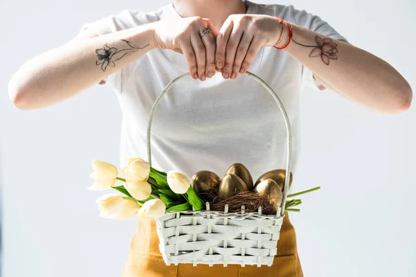 Vue recadrée d'une femme tenant un panier de paille avec des œufs de Pâques dorés et des tulipes isolées sur blanc — Photo de stock