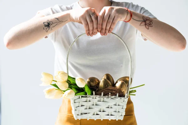 Cropped view of woman holding straw basket with golden Easter eggs and tulips isolated on white — Stock Photo