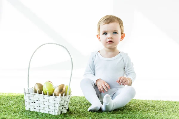 Adorável bebê sentado perto da cesta de palha com brilhantes ovos de Páscoa isolados em branco — Fotografia de Stock