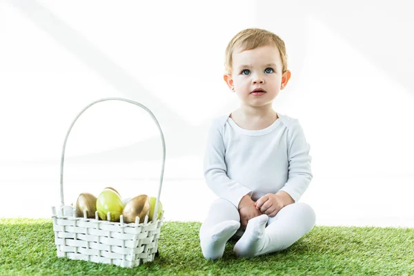Bonito criança sentado perto de palha cesta com brilhantes ovos de Páscoa isolado no branco — Fotografia de Stock