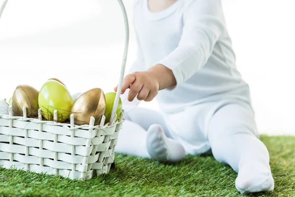 Cropped view of kid sitting near straw basket with yellow and golden chicken eggs isolated on white — Stock Photo