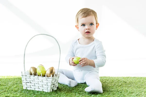 Lindo niño sosteniendo huevo de pollo amarillo mientras está sentado cerca de la canasta de paja con huevos de Pascua aislados en blanco - foto de stock