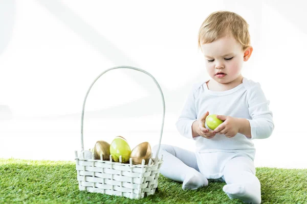 Adorable blonde child holding yellow chicken egg while sitting near straw basket with Easter eggs isolated on white — Stock Photo