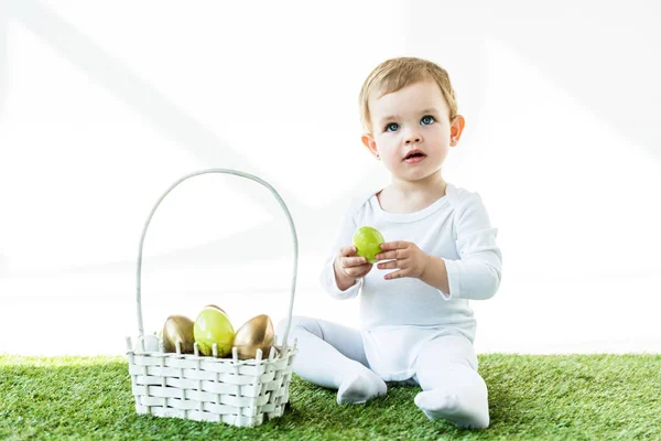 Niño rubio soñador sosteniendo huevo de pollo amarillo mientras está sentado en hierba verde cerca de la canasta de paja con huevos de Pascua aislados en blanco - foto de stock