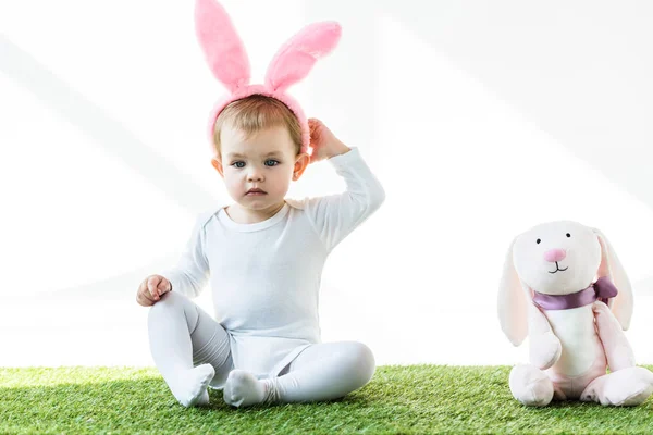 Criança adorável em orelhas de coelho headband sentado perto de coelho brinquedo engraçado isolado no branco — Fotografia de Stock