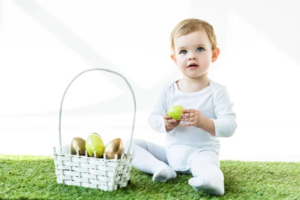Adorable kid holding yellow chicken egg while sitting on green grass near straw basket with Easter eggs isolated on white — Stock Photo