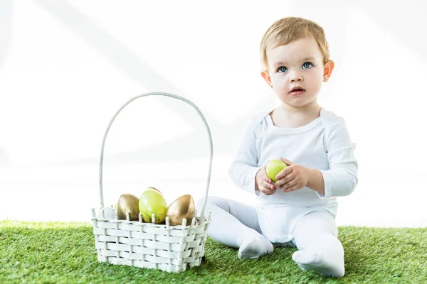 Cute blonde baby holding yellow chicken egg while sitting near straw basket with Easter eggs  isolated on white — Stock Photo