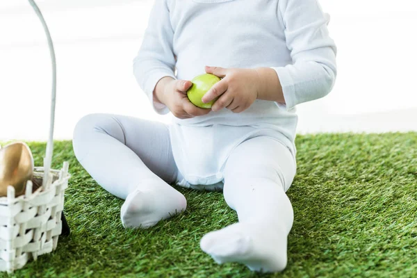 Teilansicht des Babys auf grünem Gras sitzend und gelbes Hühnerei isoliert auf weißem Grund haltend — Stockfoto