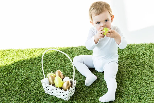 Lindo niño sosteniendo huevo de cocina amarillo cerca de la cara mientras está sentado cerca de la canasta de paja con huevos de Pascua aislados en blanco - foto de stock