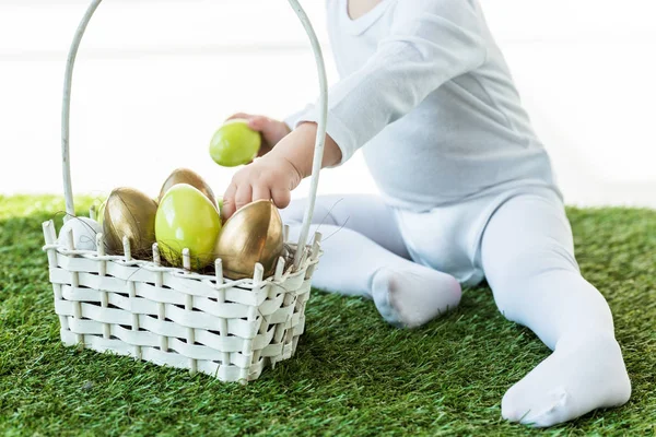 Vista parziale del bambino che prende uova di Pasqua colorate dal cestino di paglia isolato su bianco — Foto stock