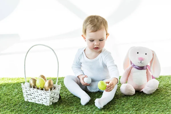 Adorable child holding colorful chicken eggs while sitting near straw basket and toy rabbit isolated on white — Stock Photo