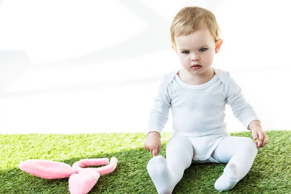 Mignon enfant assis sur herbe verte près des oreilles de lapin bandeau isolé sur blanc — Photo de stock