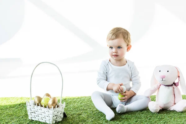 Cute baby holding colorful chicken eggs while sitting near straw basket and toy rabbit isolated on white — Stock Photo