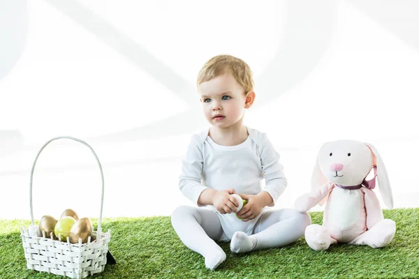 Adorable niño sentado cerca de canasta de paja con huevos de Pascua y conejo de juguete aislado en blanco - foto de stock
