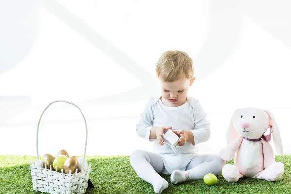 Cute child holding white box while sitting on green grass near toy rabbit and straw basket with Easter eggs isolated on white — Stock Photo