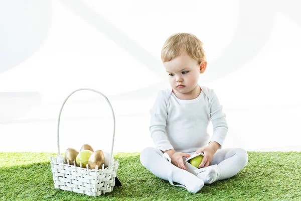 Adorable child sitting on green grass near yellow and golden Easter eggs isolated on white — Stock Photo