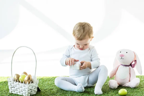 Adorable kid holding white box while sitting on green grass near toy rabbit and straw basket with Easter eggs isolated on white — Stock Photo