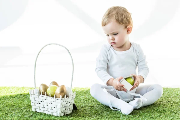 Adorable kid holding box with yellow egg while sitting near straw basket with Easter eggs isolated on white — Stock Photo