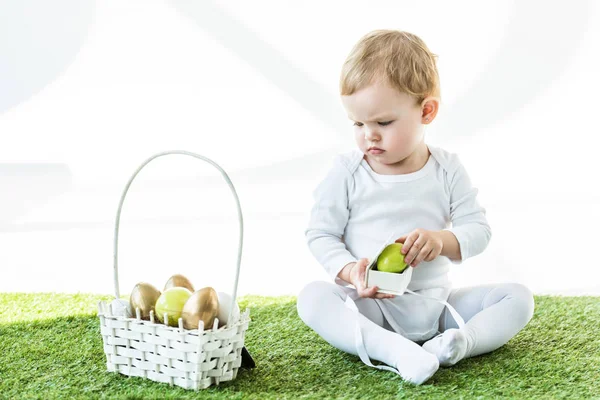 Mignon enfant tenant boîte avec oeuf jaune et regardant panier de paille avec oeufs de Pâques isolé sur blanc — Photo de stock