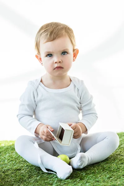 Entzückendes Kind sitzt auf grünem Gras mit weißem Karton und gelbem Hühnerei isoliert auf weißem — Stockfoto