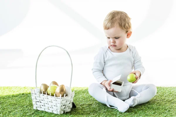 Cute kid holding white box and yellow chicken egg while sitting near straw basket with Easter eggs isolated on white — Stock Photo