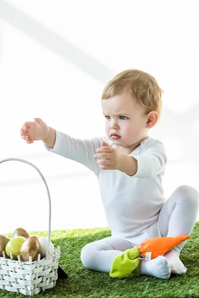 Niedliches Kind mit ausgestreckten Händen sitzt neben Strohkorb mit bunten Ostereiern isoliert auf weiß — Stockfoto