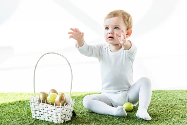 Adorable baby with outstretched hands sitting near straw basket with colorful Easter eggs isolated on white — Stock Photo