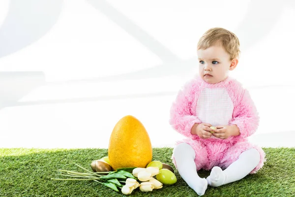 Cute baby in pink fluffy costume sitting near yellow ostrich egg, colorful chicken eggs and tulips isolated on white — Stock Photo