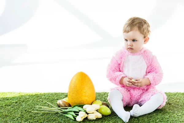 Entzückendes Kind im rosafarbenen flauschigen Kostüm, das neben gelben Straußeneiern sitzt, bunten Hühnereiern und Tulpen isoliert auf weißem Grund — Stockfoto