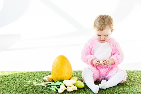 Adorable child in pink fluffy costume sitting near yellow ostrich egg, colorful chicken eggs and tulips isolated on white — Stock Photo