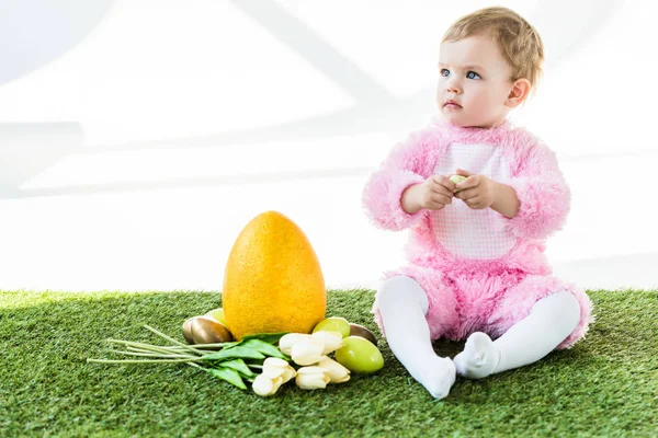 Carino bambino in costume rosa soffice seduto vicino all'uovo di struzzo giallo, uova di pollo colorate e tulipani isolati su bianco — Foto stock