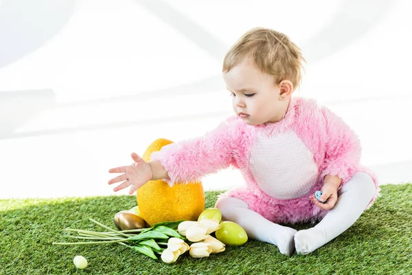 Entzückendes Kind im rosafarbenen flauschigen Kostüm sitzt neben gelben Straußeneiern, bunten Hühnereiern und Tulpen isoliert auf weißen — Stockfoto