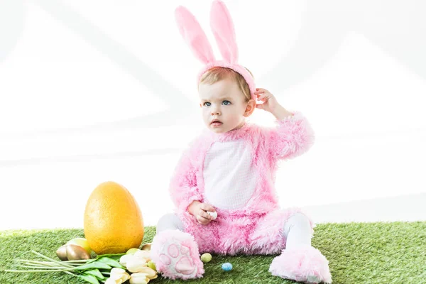 Niedliches Baby im lustigen Hasenkostüm sitzt in der Nähe von Ostereiern, Tulpen und gelben Straußeneiern isoliert auf weiß — Stockfoto