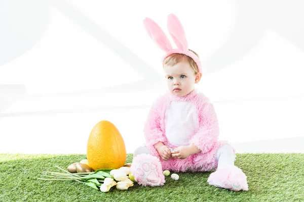 Niedliches Baby im lustigen Hasenkostüm sitzt neben gelben Straußeneiern, bunten Ostereiern und Tulpen isoliert auf weiß — Stockfoto