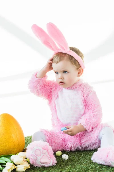 Adorable niño en divertido traje de conejo sentado cerca de huevo de avestruz amarillo, coloridos huevos de Pascua y tulipanes aislados en blanco - foto de stock