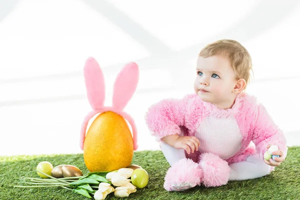 Adorable niño en traje mullido rosa sentado cerca de huevo de avestruz amarillo con diadema de orejas de conejo, coloridos huevos de Pascua y tulipanes aislados en blanco - foto de stock