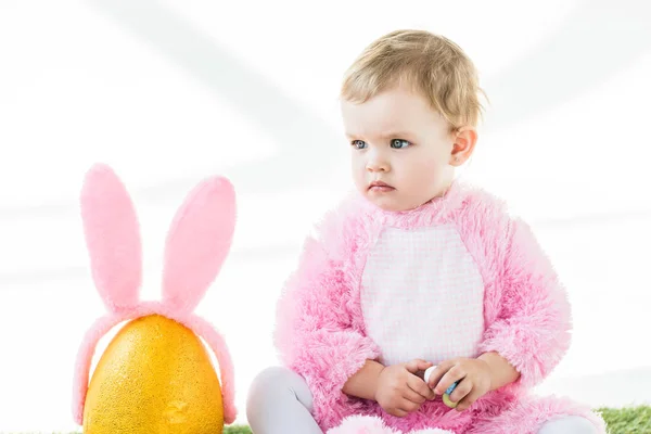 Garoto bonito em traje fofo rosa segurando ovos de codorna coloridos enquanto sentado perto de ovo de avestruz amarelo com orelhas de coelho headband isolado no branco — Fotografia de Stock