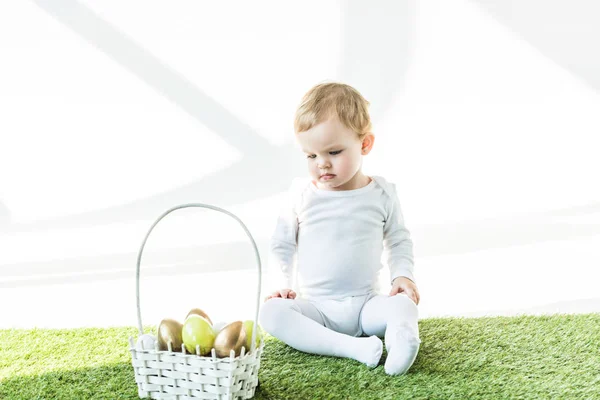 Criança bonito sentado na grama verde perto da cesta de palha com ovos de Páscoa coloridos isolados no branco — Fotografia de Stock