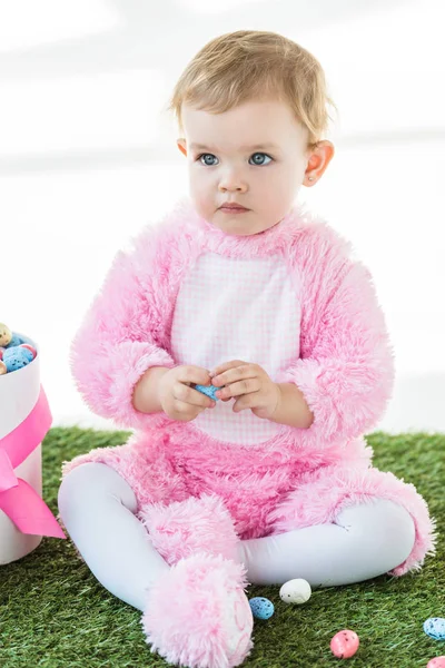 Mignon enfant rêveur en costume rose moelleux assis près de la boîte avec des œufs de Pâques isolé sur blanc — Photo de stock