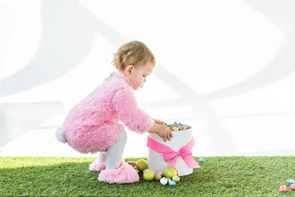 Niedliches Kind im rosafarbenen flauschigen Kostüm steht neben Schachtel mit rosa Schleife und bunten Ostereiern isoliert auf weiß — Stockfoto