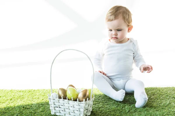 Adorable child sitting on green grass near straw basket with yellow and golden Easter eggs isolated on white — Stock Photo