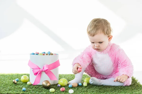 Niedliches Baby im rosafarbenen flauschigen Kostüm sitzt auf grünem Gras in der Nähe von Geschenkbox mit rosa Schleife und bunten Ostereiern isoliert auf weiß — Stockfoto