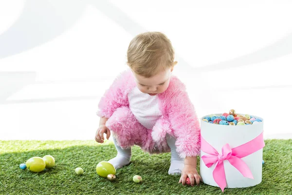 Entzückendes Kind im rosafarbenen flauschigen Kostüm, das bunte Eier aus grünem Gras in der Nähe von Geschenkschachtel mit rosa Schleife isoliert auf weiß nimmt — Stockfoto