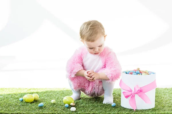 Niedliches Baby im rosafarbenen flauschigen Kostüm sitzt auf grünem Gras in der Nähe von Geschenkbox mit rosa Schleife und bunten Ostereiern isoliert auf weiß — Stockfoto